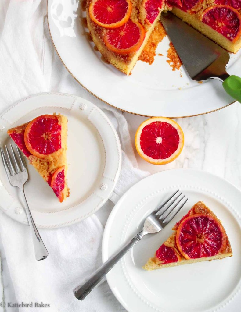 Two slices of blood orange olive oil upside down cake on a white background with the rest of the cake next to them