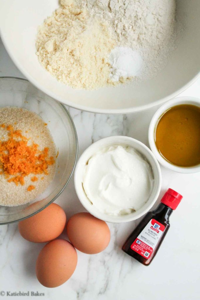 ingredients for the cake on a white marble background: flour, sugar, orange zest, olive oil, yogurt, vanilla extract, eggs