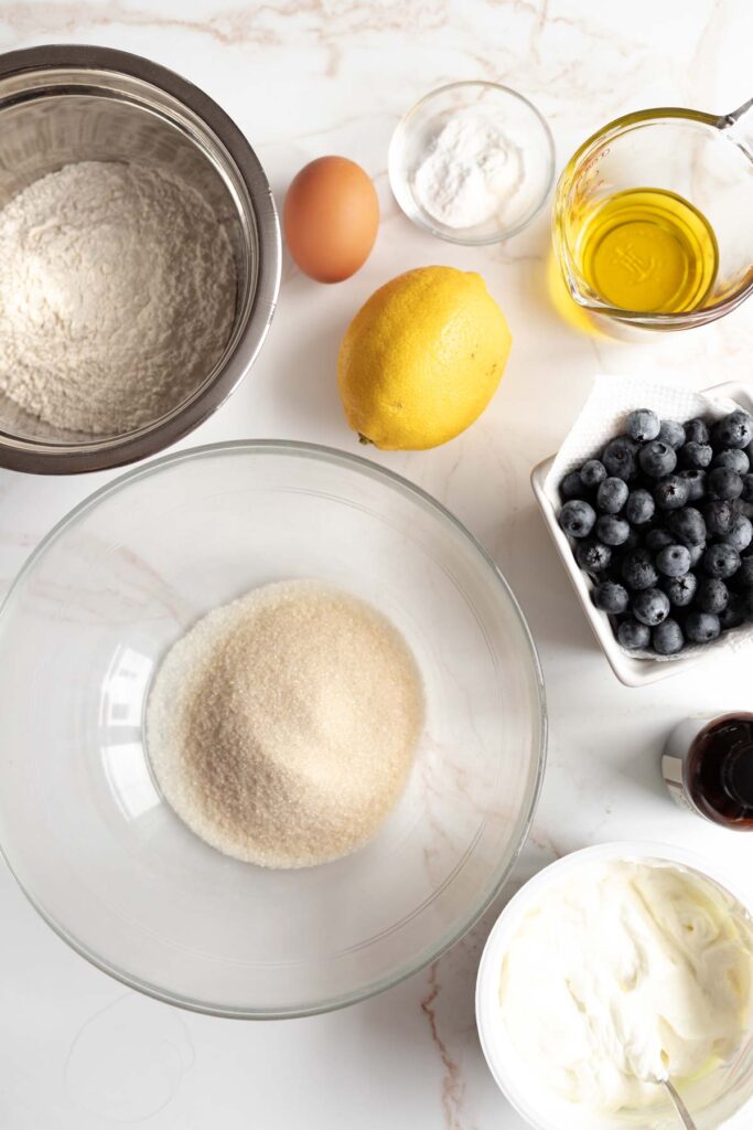 Overhead view of the ingredients needed for the muffins in varying sizes of bowls, set on a white marble background.