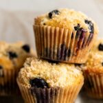 two lemon blueberry muffins made with greek yogurt stacked on top of each other in front of a cream colored background, with more muffins behind