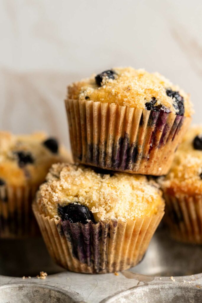 two lemon blueberry muffins made with greek yogurt stacked on top of each other in front of a cream colored background, with more muffins behind