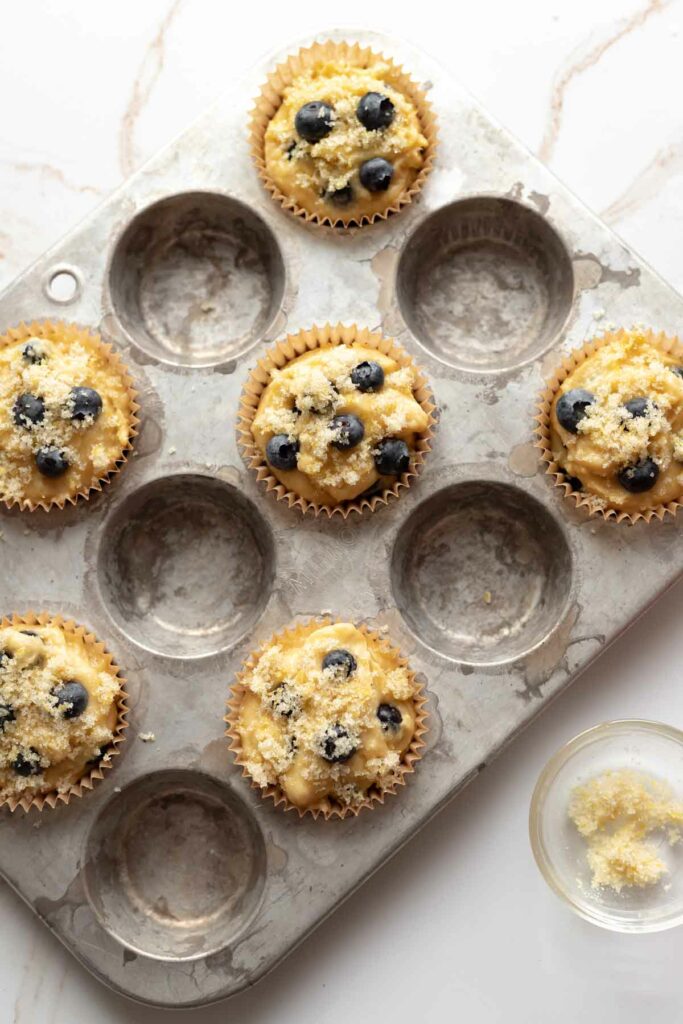 overhead view of muffin batter scooped into in every other cup of a 12 cup muffin tin, with additional blueberries and lemon sugar placed on top of each