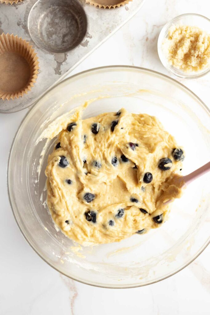 overhead view of the blueberry muffin batter in a large glass bowl resting, to be scooped into the muffin tin next to it