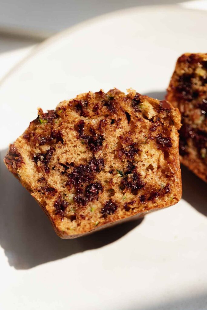 a zucchini muffin on a white plate, cut in half to show the distribution of mini chocolate chips throughout the muffin