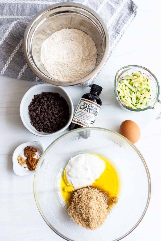 Ingredients for the recipe arranged on a white surface: shredded zucchini, flour, olive oil, brown sugar, greek yogurt, egg, vanilla extract, cinnamon, nutmeg, ginger, mini chocolate chips