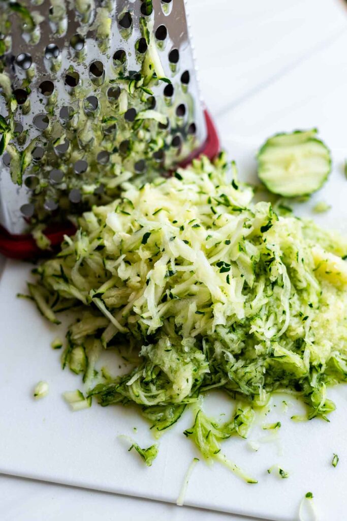 shredded zucchini next to a box grater on a white cutting board