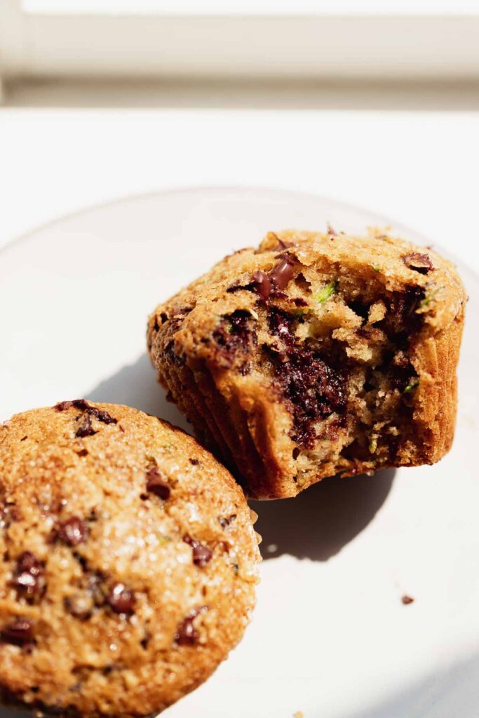 a zucchini chocolate chip muffin on a white plate with a bite taken out of it, showing the bits of shredded zucchini and melty chocolate inside